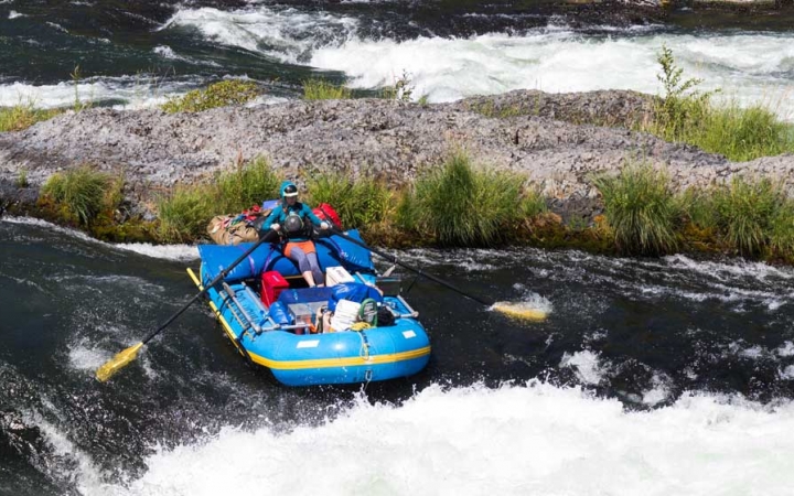 A person paddles a packed raft through whitewater in front of a rocky shore.
